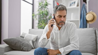 Genç İspanyol beyaz saçlı adam telefonda konuşuyor. Evdeki kanepede oturuyor.