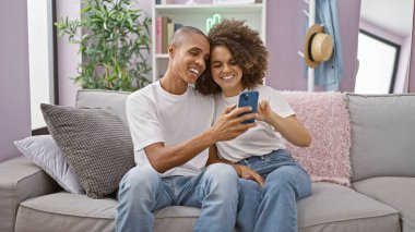 Güzel bir çift, mutlu bir şekilde evlerindeki koltuklarında oturup, mutlu mesut mesajlaşırken, gülen yüz ifadelerinden neşe saçıyor.