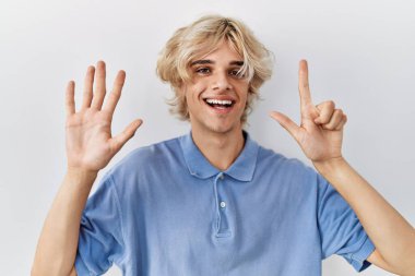 Genç modern adam izole edilmiş arka planda duruyor ve yedinci parmaklarıyla işaret ederken kendinden emin ve mutlu bir şekilde gülümsüyor.. 
