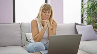 Konforlu evinde, ciddi ve odaklanmış orta yaşlı sarışın bir kadın koltukta oturuyor, tamamen içine kapanık, oturma odasındaki bilgisayarını kullanıyor..
