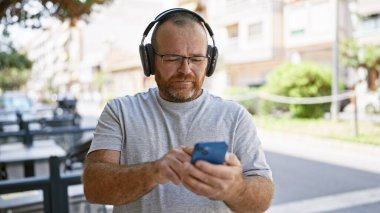 Beyaz adam sokakta akıllı telefon kullanıyor. Kulaklık takıyor.