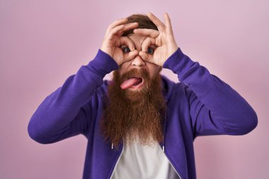 Pembe arka planda uzun sakallı beyaz bir adam dürbün gibi jest yapıyor. Dürbün gibi dilini çıkarıyor, gözler parmaklarından bakıyor. çılgın ifade. 