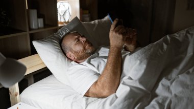 Yakışıklı, orta yaşlı, beyaz sakallı, yatakta dinlenen, akıllı telefonunu başucundaki lambada kullanan bir adam. Yatak odasının rahat sınırları içinde dijital dünyada kayboldu..