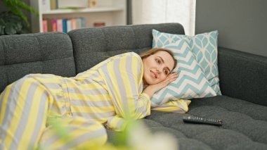 Young blonde woman watching tv lying on sofa at home