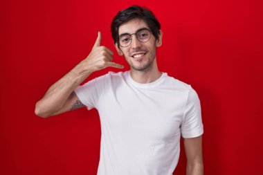 Young hispanic man standing over red background smiling doing phone gesture with hand and fingers like talking on the telephone. communicating concepts. 