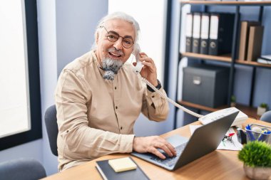 Middle age grey-haired man business worker talking on telephone using laptop at office