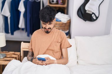 Genç İspanyol adam yatakta akıllı telefon kullanıyor.