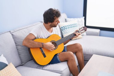 Young hispanic man playing classical guitar sitting on sofa at home