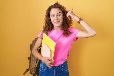 Sırt çantası takan, elinde kitaplarla gülümseyen beyaz bir kadın. Tek parmakla işaret ediyor. Harika bir fikir ya da düşünce. 