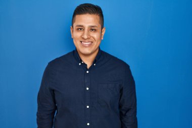 Yüzünde mutlu ve havalı bir gülümsemeyle mavi arka planda duran İspanyol genç adam. şanslı kişi. 