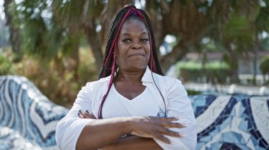 Afro-Amerikan kadın, kollarını kavuşturup, güzel, rahat bir gülümseme takınarak, neşeyle şehir parkında duruyor.