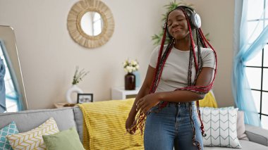 Neşeli Afro-Amerikan kadın rock 'çı, müzik dinleyen, salonda özgüvenle dans eden, evinde rahatlatıcı yaşam tarzının keyfini çıkarırken gülümseyen