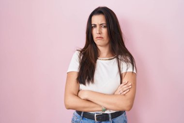 Pembe arka planda duran esmer kadın şüpheci ve gergin, yüzünde çapraz kollu tasvip etmeyen ifade. negatif kişi. 