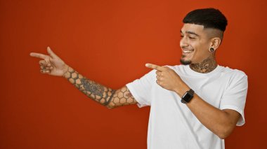 Neşeli genç latin adam kendinden emin, kendine güvenen, enfeksiyon derecesinde mutlu bir gülümseme sergilerken dövmeli ellerini göstererek, canlı, izole bir kırmızı arkaplanı işaret ediyor..