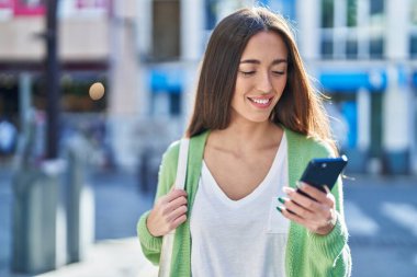 Genç, güzel, İspanyol bir kadın sokakta akıllı telefon kullanarak gülümsüyor.