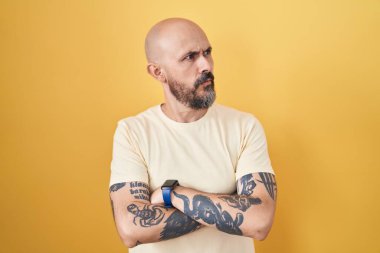 Sarı arka planda duran İspanyol dövmeli adam kollarını kavuşturmuş, ikna olmuş ve kendinden emin bir şekilde yan tarafa bakıyor. 