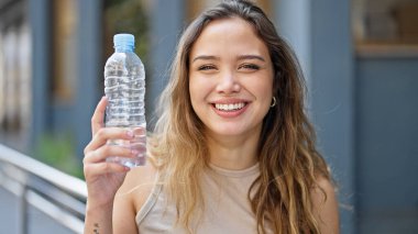 Genç, güzel, İspanyol bir kadın elinde bir şişe suyla sokağa gülümsüyor.
