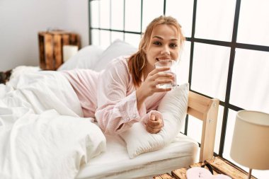 Beyaz kadın yatak odasında uzanmış bir bardak su içiyor.