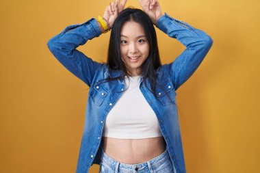 Sarı arka planda duran genç Asyalı kadın komik ve çılgın pozlar veriyor parmakları kafasında tavşan kulağı, neşeli bir şekilde gülümsüyor 