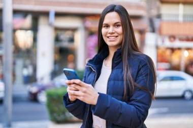 Genç, güzel, İspanyol bir kadın sokakta akıllı telefon kullanarak gülümsüyor.