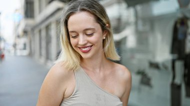 Genç sarışın kadın gülümsüyor. Kendinden emin bir şekilde sokağa bakıyor.