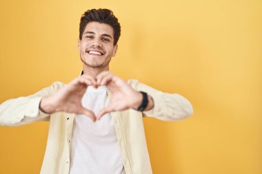 Sarı arka planda dikilen genç İspanyol aşık gülümseyen ve elleriyle kalp sembolü yapan adam. romantik kavram. 