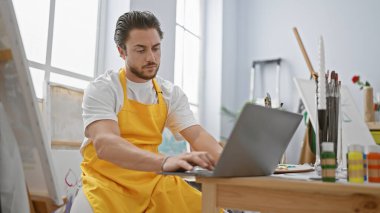 Genç İspanyol ressam, sanat stüdyosunda ciddi yüzlü dizüstü bilgisayar kullanıyor.