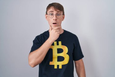 Bitcoin tişört giyen beyaz, sarışın adam elleri çenesinde, inanamama, şaşkınlık ve şaşkınlık dolu bir ifade takınıyor. 