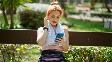 Parktaki bankta oturan akıllı telefon ve kulaklık kullanan genç kızıl saçlı kadın.