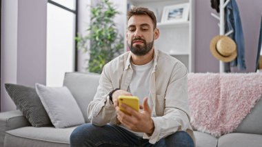 Yakışıklı, sakallı, İspanyol asıllı, ciddi mesajlaşmalara kendini kaptırmış. Oturma odasındaki kanepede rahatça oturup akıllı telefon teknolojisi kullanıyor.