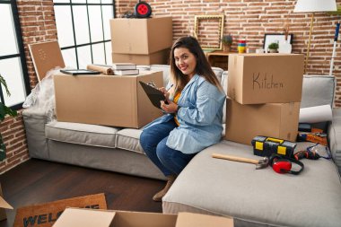 Young beautiful plus size woman writing on document sitting on sofa at new home