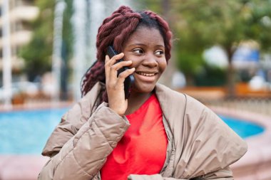 Afro-Amerikalı kadın gülümsüyor. Parktaki akıllı telefondan konuşuyor.