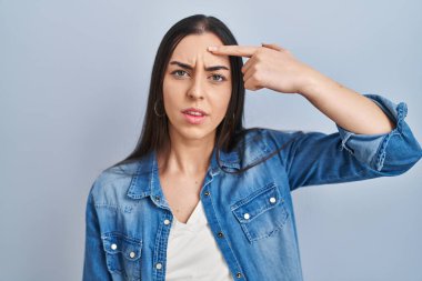 Mavi arka planda dikilen İspanyol kadın alnındaki sivilceden mutsuz olduğunu gösteriyor, siyah nokta enfeksiyonu. sivilce ve deri problemi 