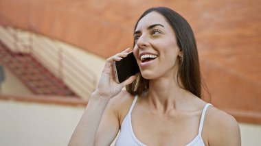 Genç, güzel, İspanyol bir kadın gülümsüyor ve caddede akıllı telefondan konuşuyor.