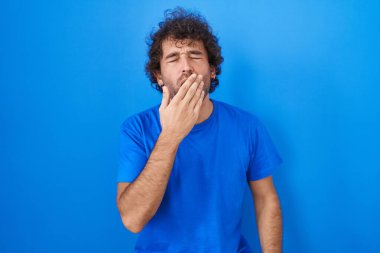 Mavi arka planda dikilen Latin asıllı genç adam elleriyle ağzını kapatmaktan sıkılmış. Huzursuz ve uykulu.. 
