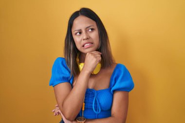 Sarı arka planda dikilen Latin kökenli genç bir kadın bir soru hakkında endişeli ve gergin bir şekilde düşünüyor. 