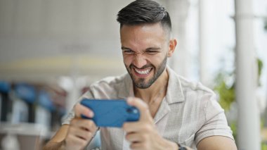 Latin asıllı genç adam, kahvecinin terasında video oyunu oynarken kendine güveniyor.