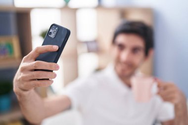 Young hispanic man making selfie by smartphone drinking coffee at home