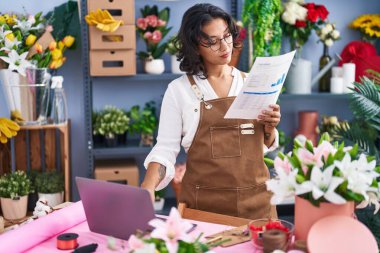 Young beautiful hispanic woman florist using laptop reading document at flower shop