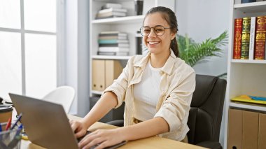 Genç, güzel İspanyol kadın iş sihrini bir dizüstü bilgisayarla yapıyor. Profesyonel bir ofis içinde neşe ve başarı yayıyor..