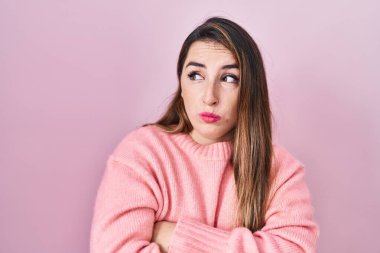 Young hispanic woman standing over pink background looking to the side with arms crossed convinced and confident 