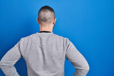 Mavi arka planda dikilen İspanyol adam kolları vücudunda geriye bakıyor. 