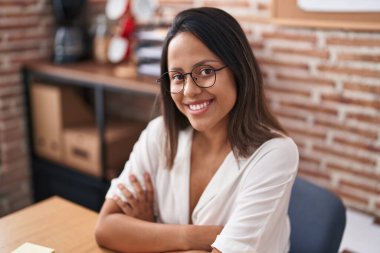 Young hispanic woman business worker using laptop sitting with arms crossed gesture at office