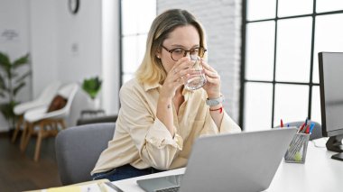 Genç sarışın iş kadını ofiste laptopla su içiyor.