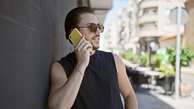 Güneş gözlüğü takan genç bir İspanyol sokakta akıllı telefondan konuşuyor.