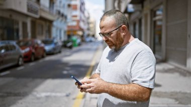 Beyaz adam sokakta ciddi bir ifadeyle akıllı telefon kullanıyor.