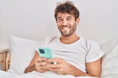 Young man using smartphone sitting on bed at bedroom