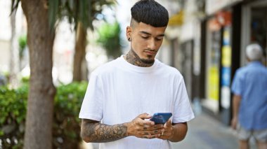 Havalı ve dövmeli genç latin adam akıllı telefonuna odaklanmış güneşli bir şehir caddesinde dikilirken mesaj atıyor ciddi bir ifade ve rahat bir yaşam tarzı sergiliyor.
