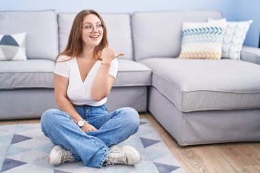 Oturma odasında yerde oturan, gülümseyen, gülümseyen ve baş parmağıyla yan tarafı gösteren beyaz bir kadın.. 