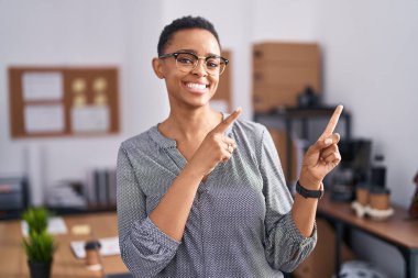 Ofiste çalışan Afro-Amerikalı kadın gözlüklü, gülümseyen ve kameraya bakıp iki elini ve parmağını yana doğru gösteren.. 